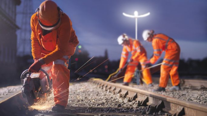 Arbeiter in orangefarbener Sicherheitskleidung reparieren Gleise bei Nacht.